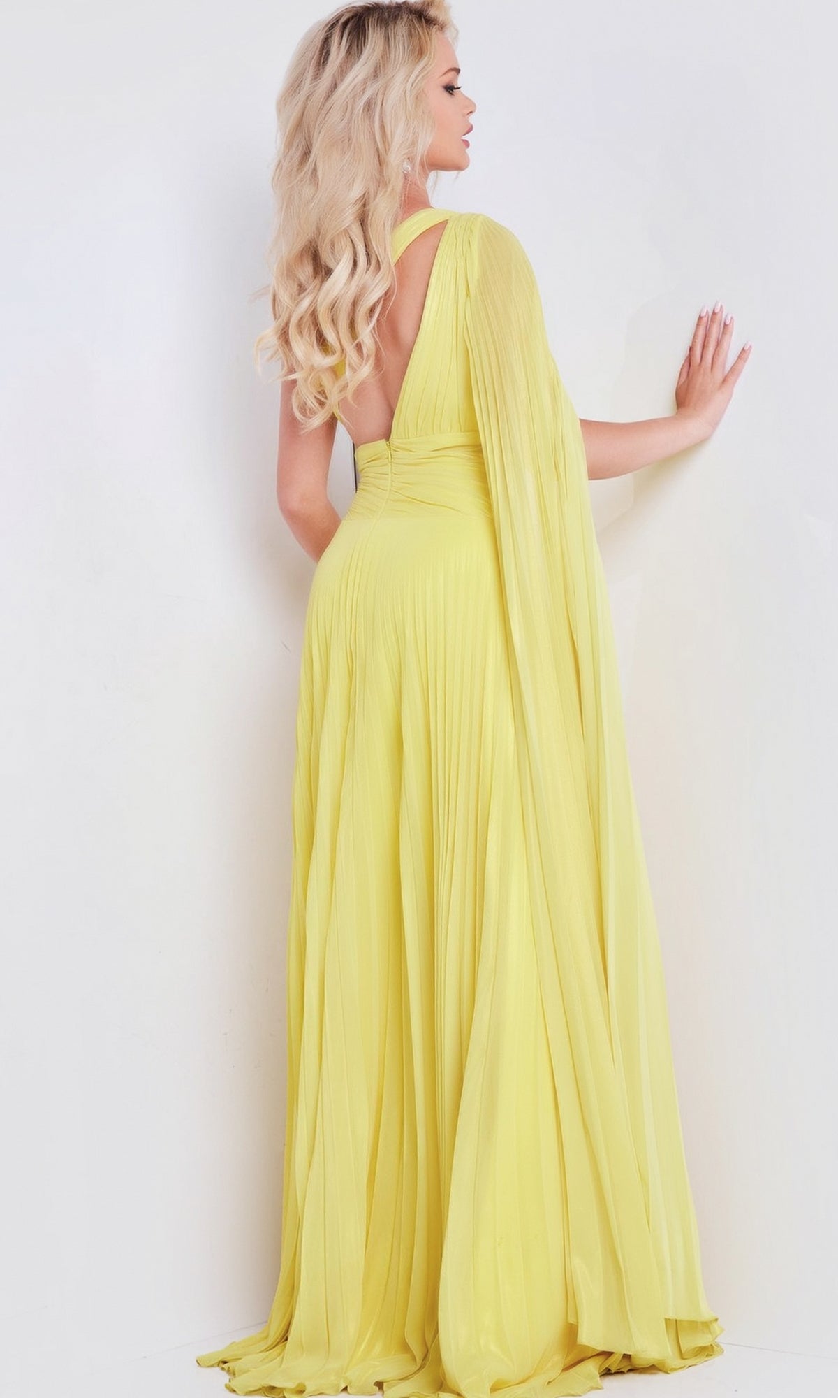 A woman with long blonde hair stands facing a white wall, wearing the Long Prom Dress 36462 by Jovani—a flowing, pleated chiffon gown—with one hand resting on the wall.